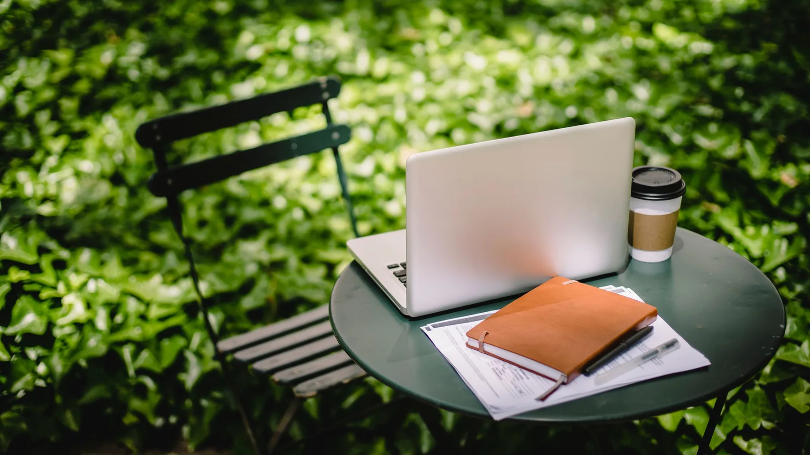 Laptop, notitieboek en koffie op een tuintafel tussen het groen